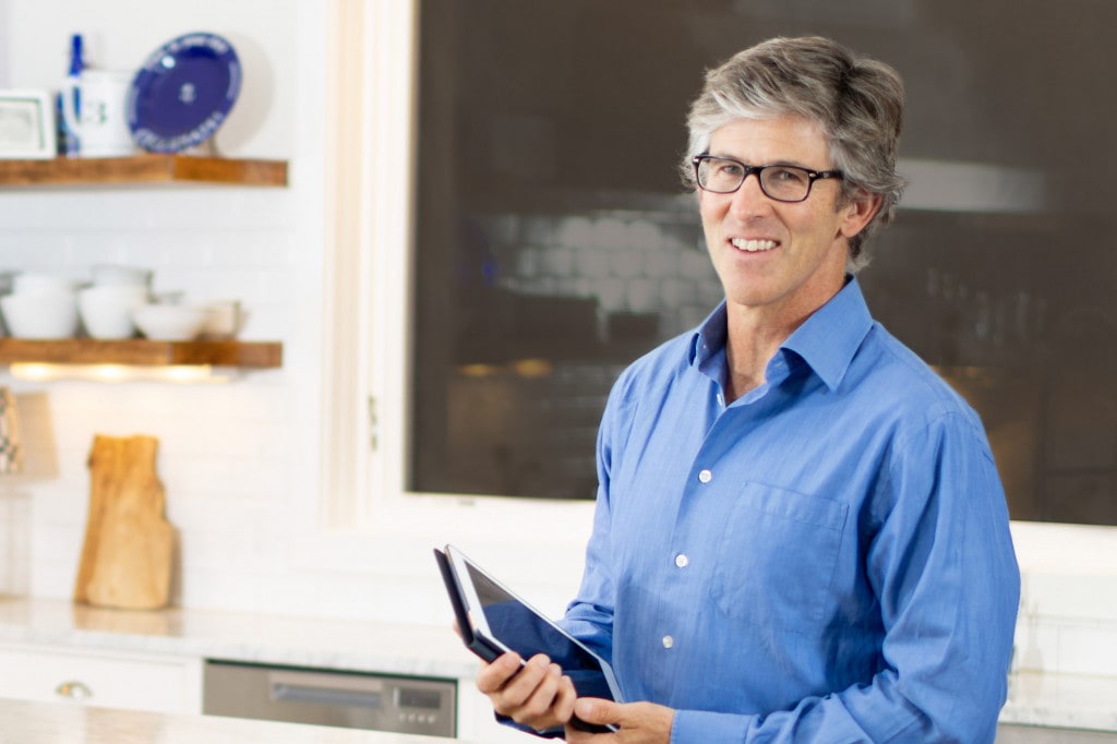 A man in glasses standing in a kitchen holding a tablet.